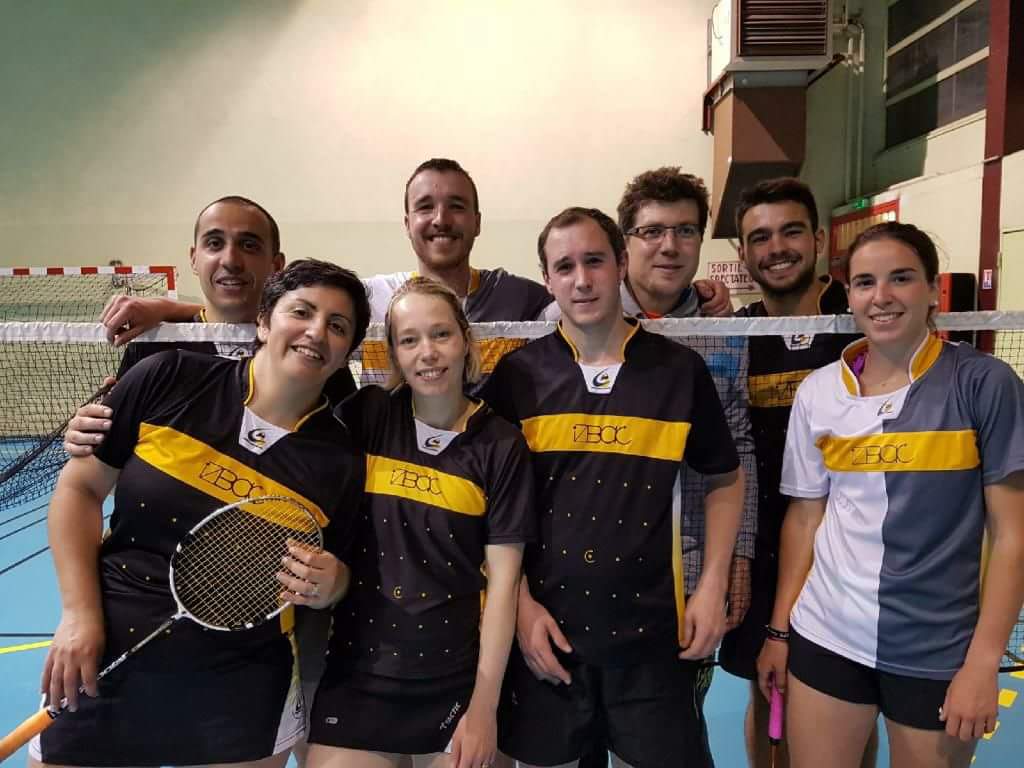 8 joueurs de badminton debout. 4 devant un filet et 4 derrière