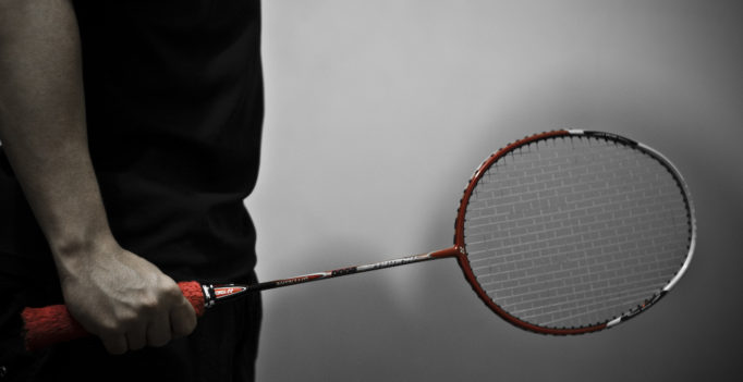 Une photo en noir et blanc en gros plan sur un homme tentant une raquette de badminton. Celle-ci est en couleur rouge